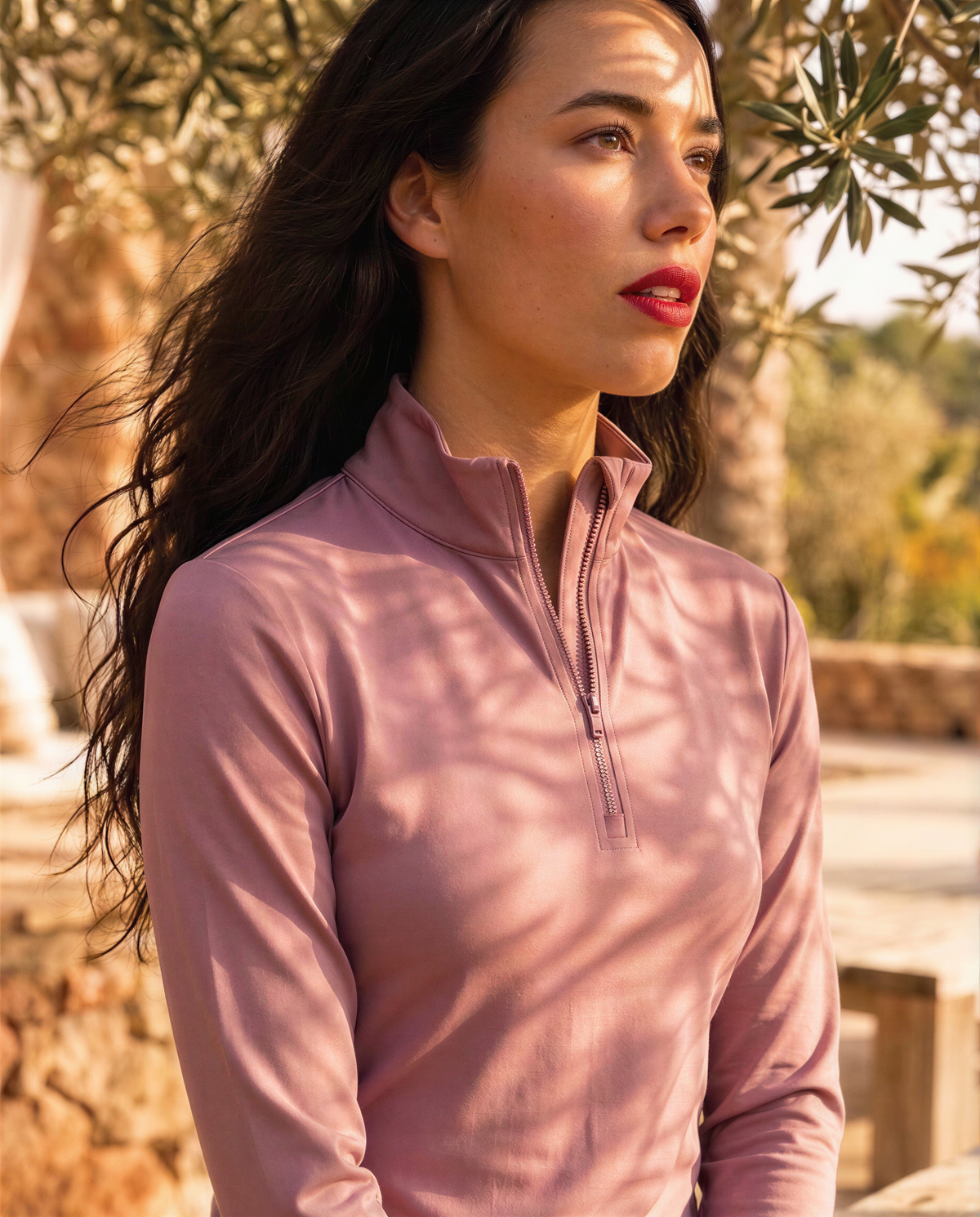 A woman wearing a dusty pink long-sleeve quarter-zip athletic top, photographed outdoors with warm sunlight and leaf shadows on her face and clothing.