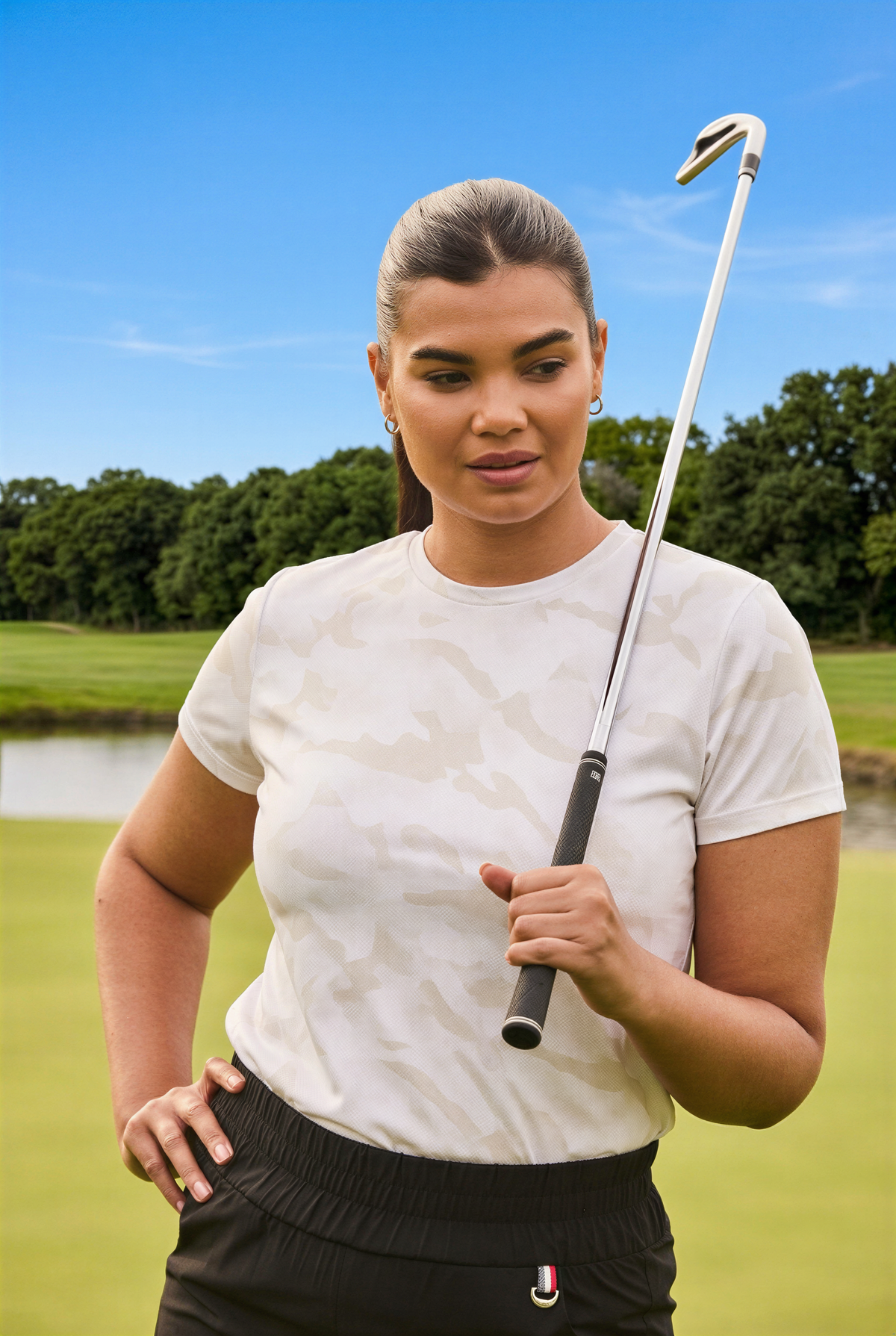 A woman on a golf course wearing a white camouflage short-sleeve t-shirt and black pants, holding a golf club.