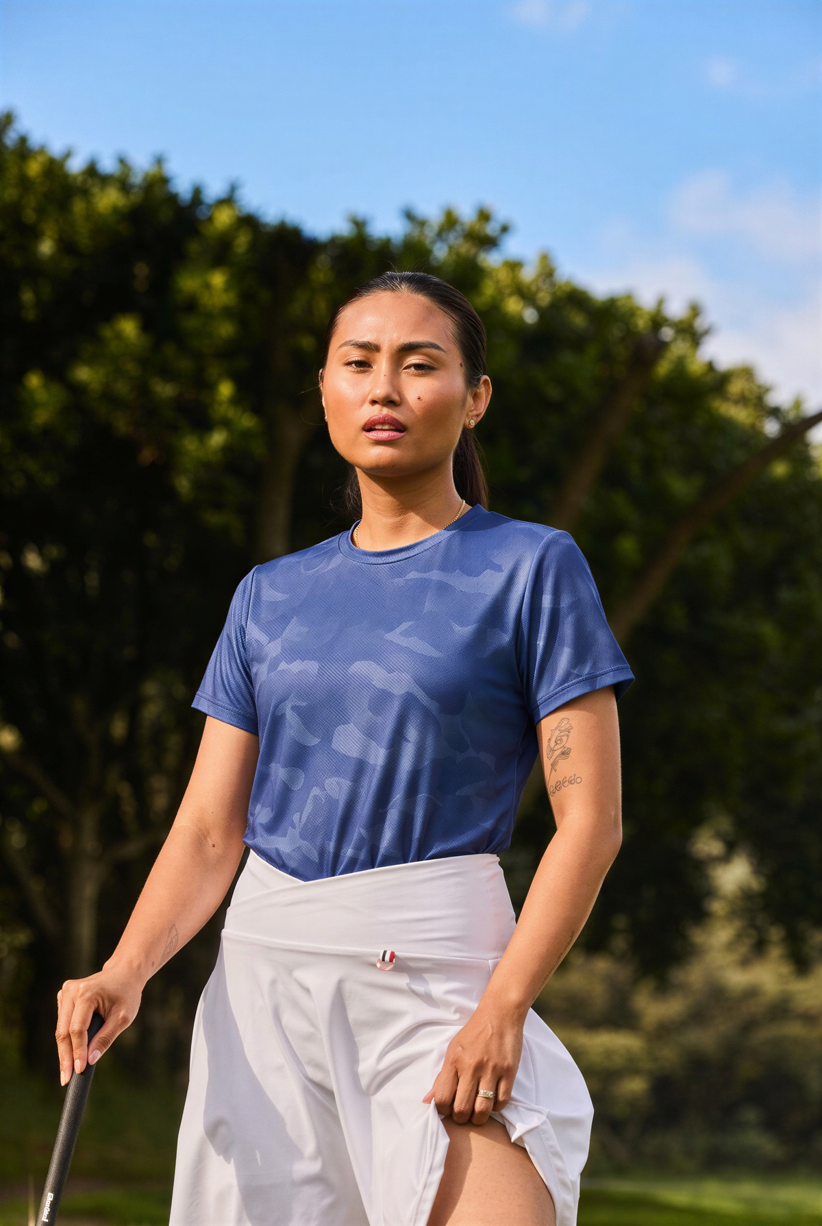 A woman standing outdoors on a golf course wearing a blue patterned short-sleeve athletic shirt and a white wrap skirt, holding a golf club.