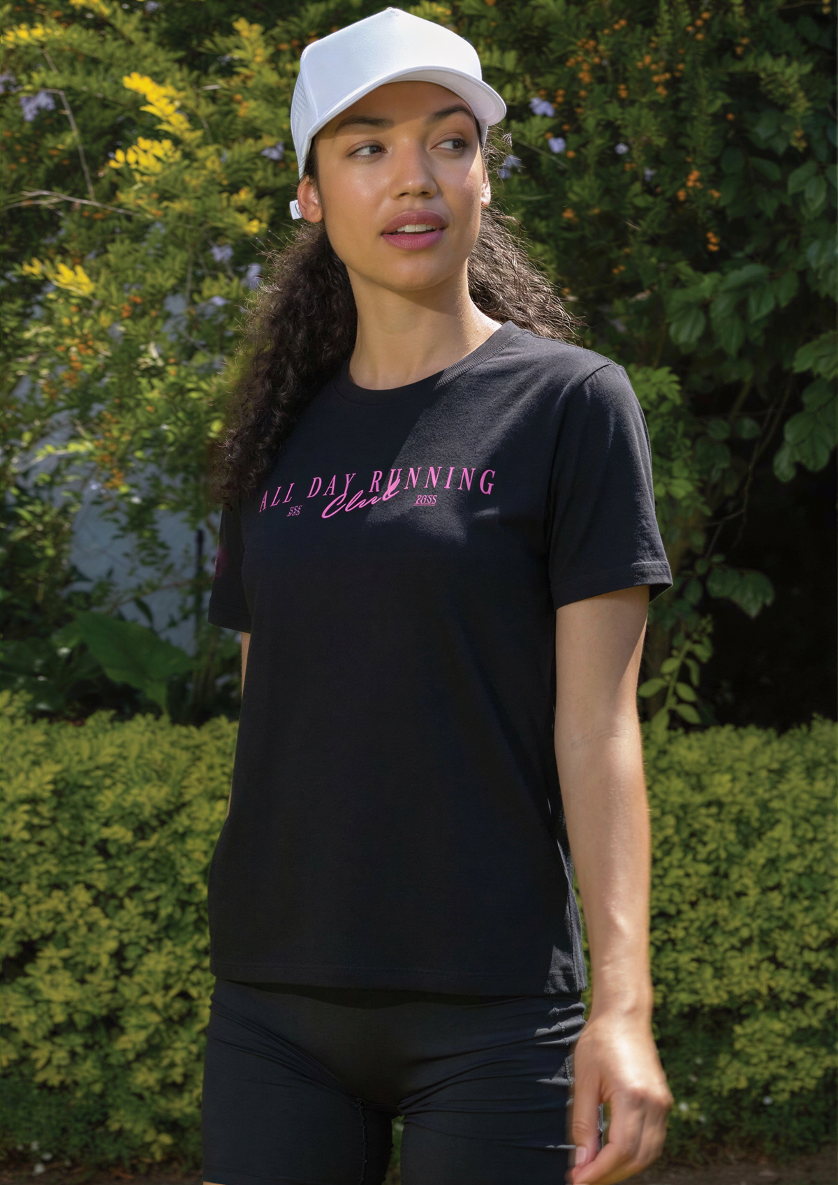 A woman wearing a black athletic t-shirt with pink text that reads 'ALL DAY RUNNING Club' and a white baseball cap, standing outdoors in front of green bushes.