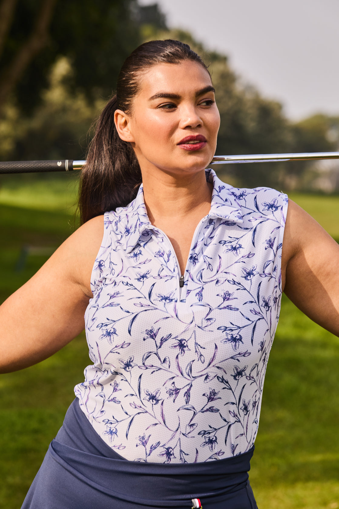 A woman on a golf course wearing a sleeveless white polo shirt with a blue floral pattern and a navy blue skirt, holding a golf club over her shoulder.