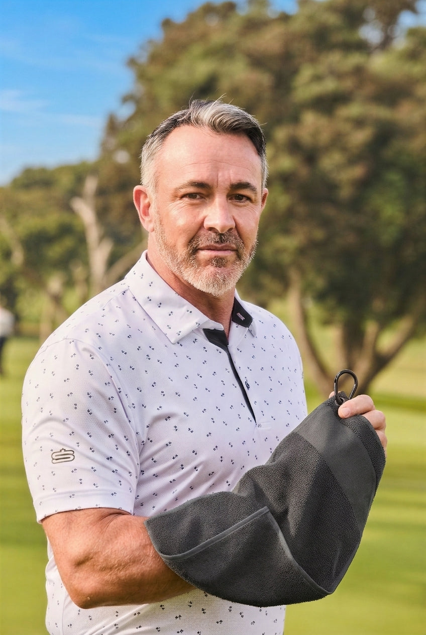 A man on a golf course wearing a white patterned polo shirt and holding a black golf towel over his arm.