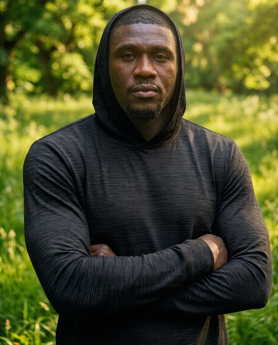 A man wearing a dark charcoal gray long-sleeve athletic hoodie with the hood up, standing outdoors in a grassy, wooded area with his arms crossed.