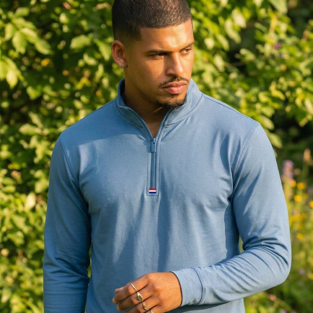 A man wearing a light blue long-sleeve quarter-zip pullover with a small red, white, and blue logo on the chest, standing outdoors in front of a green leafy background.