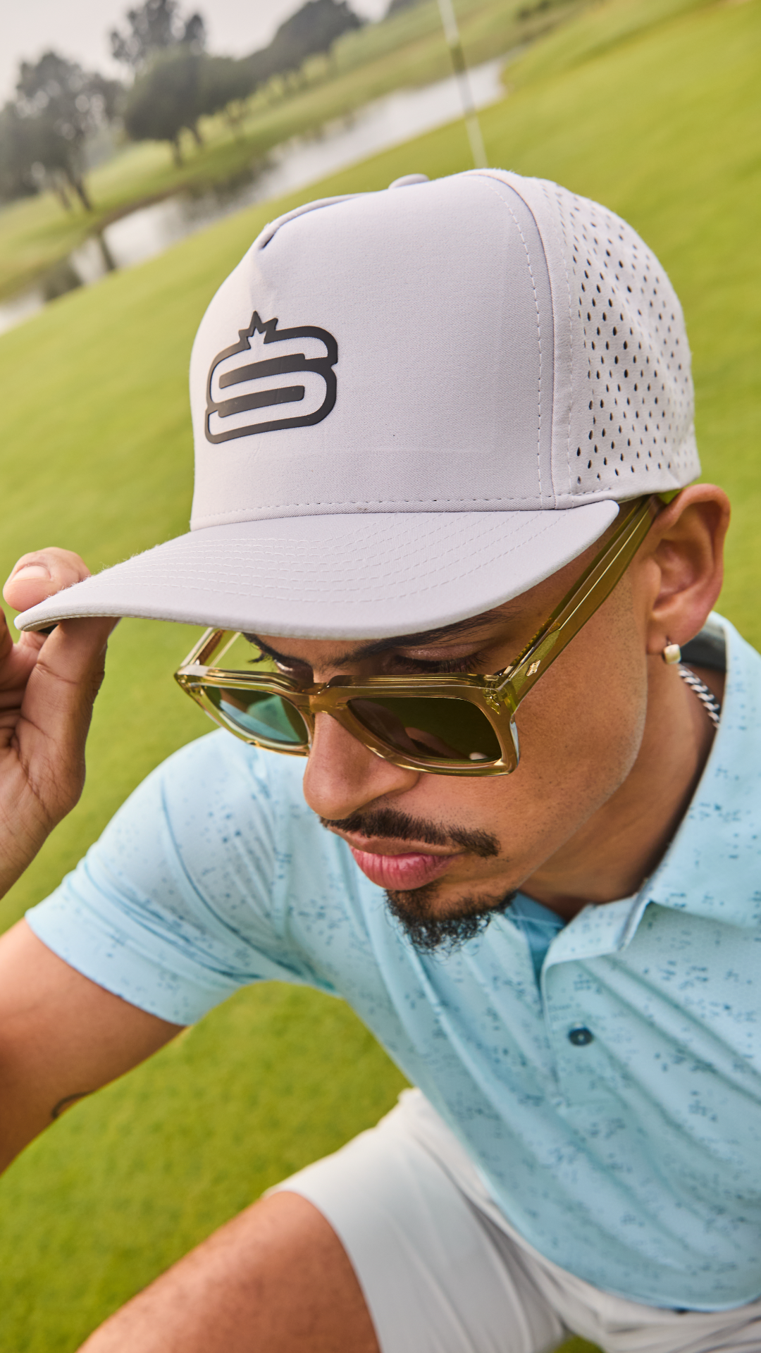 A man on a golf course wearing a white trucker-style cap with a black stylized 'S' logo, gold-framed sunglasses, and a light blue polo shirt.