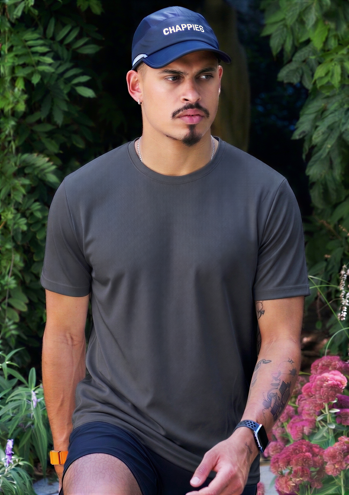 A man wearing a dark gray crewneck t-shirt, navy blue shorts, and a navy blue baseball cap with the brand name 'CHAPPIES' in white, posed outdoors in front of a lush green garden background.