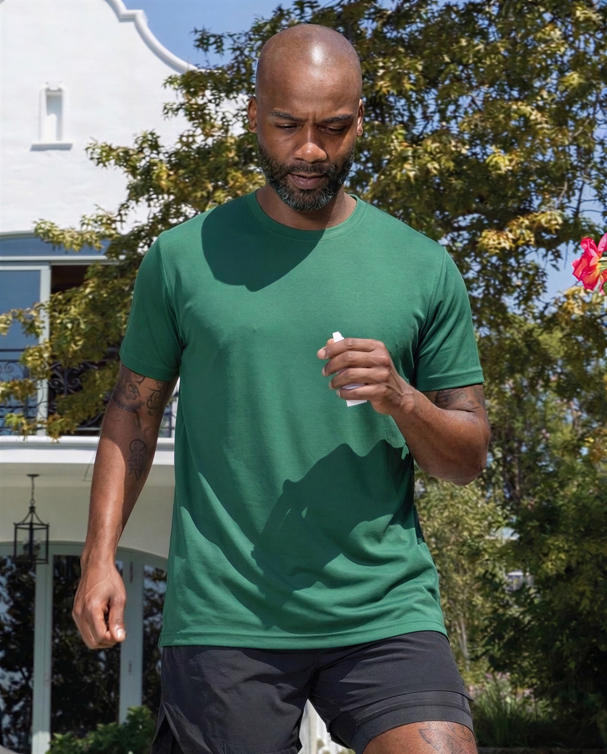 A man wearing a green short-sleeve crewneck t-shirt and black athletic shorts, standing outdoors in front of a white house and green trees.